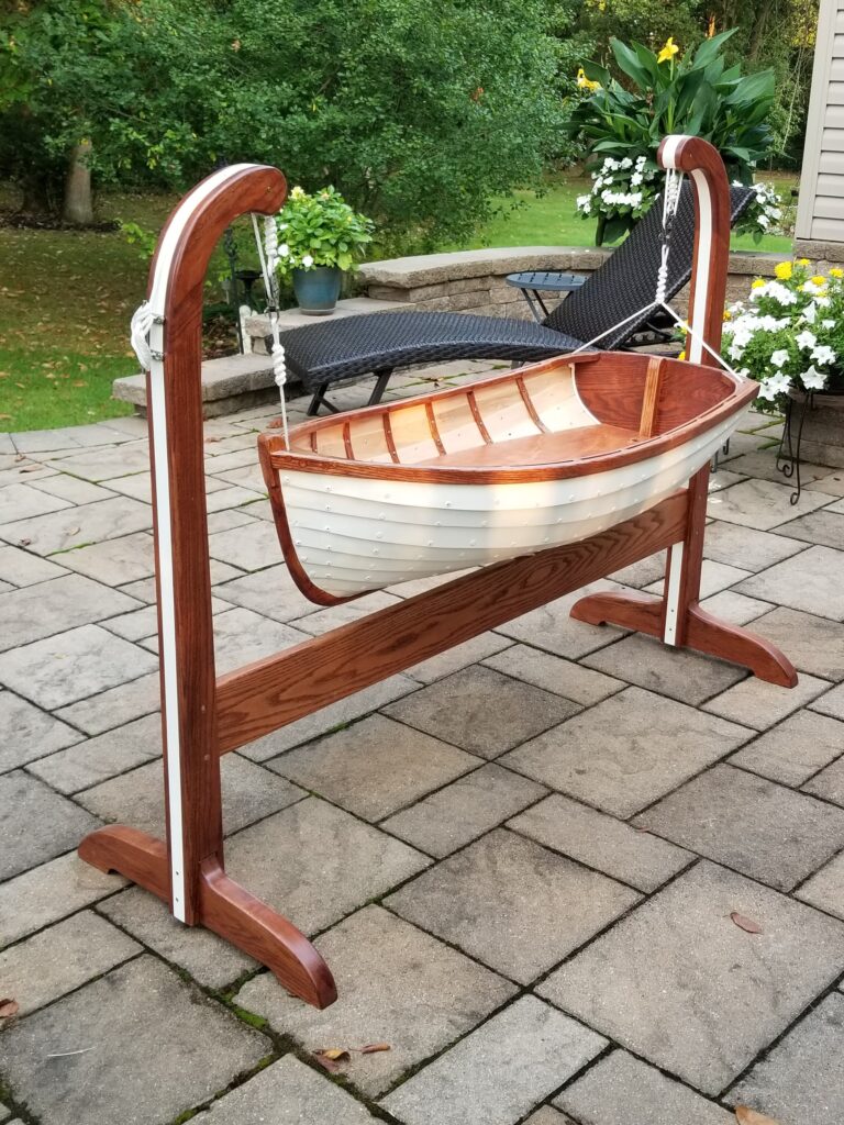 Cradle in the style of a rowboat made by Paul Musso. The cradle has a white outer hull and is hung from a frame by carefully tied ropes. The inside shows the careful craftsmanship by Paul.